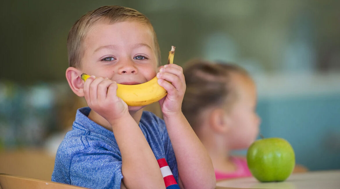 A child holding a banana in front of their face, creating a smile. Promoting healthy school snacks to protect children’s teeth.