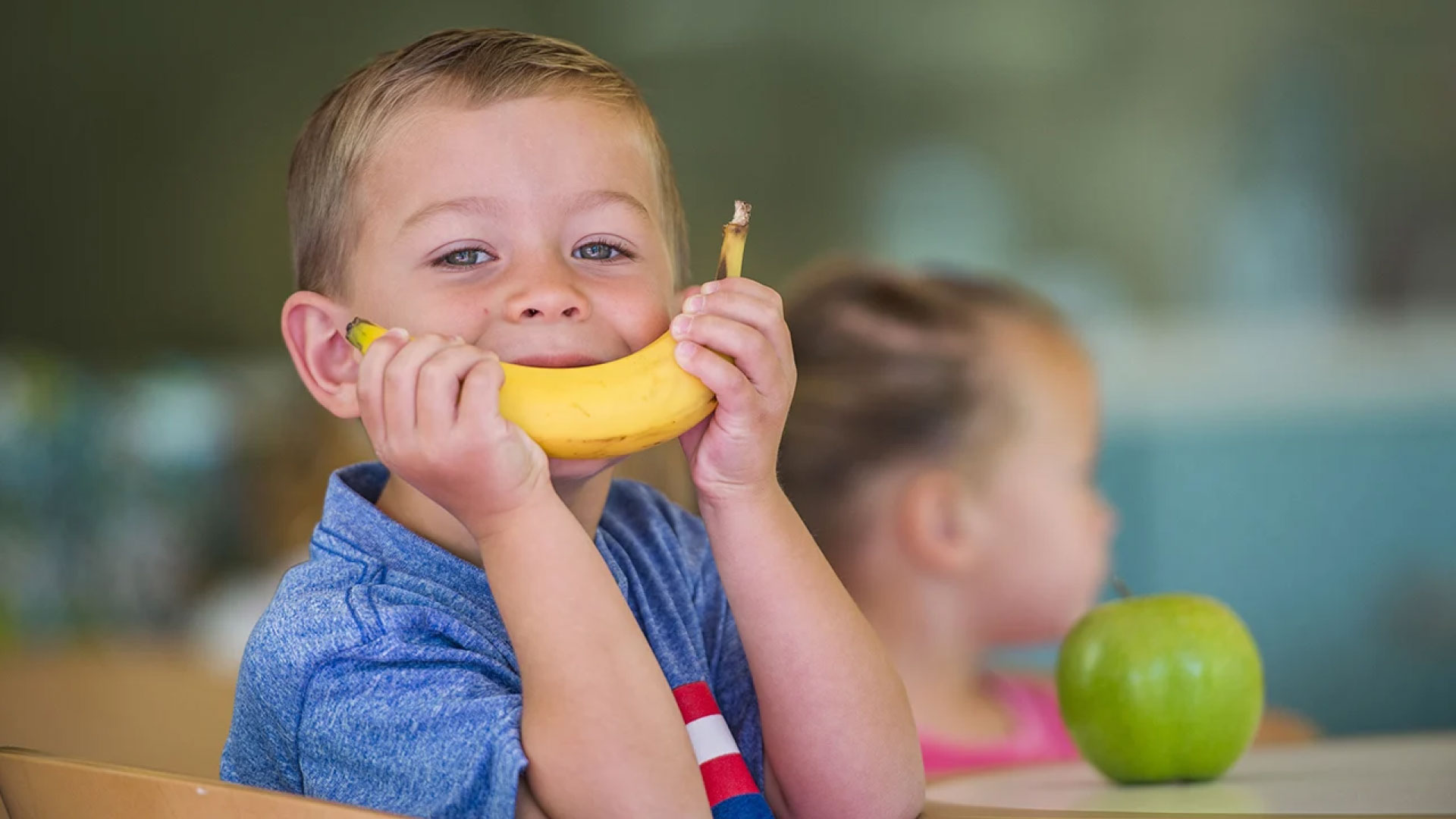 A child holding a banana in front of their face, creating a smile. Promoting healthy school snacks to protect children’s teeth.