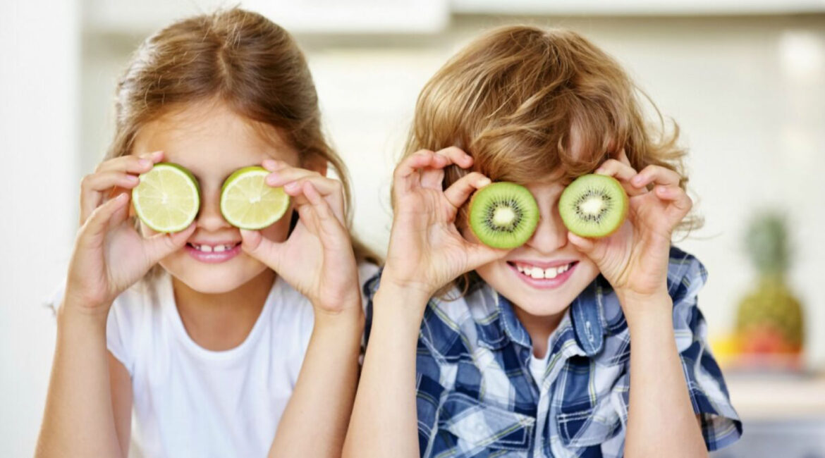 Two children hiding their eyes behind slices of orange and kiwi, representing the importance of vitamin C and calcium for healthy gums and teeth. Vitamin C and Calcium: Essential for Healthy Gums and Teeth.