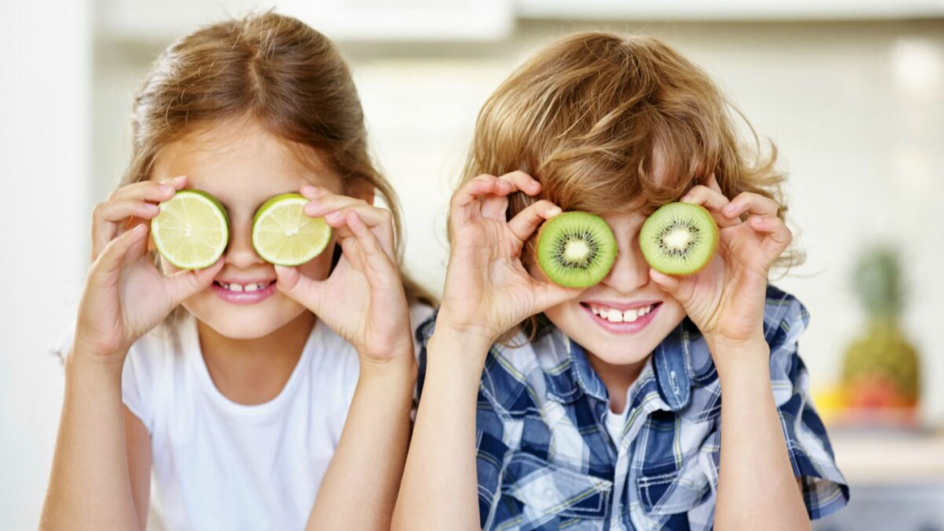 Two children hiding their eyes behind slices of orange and kiwi, representing the importance of vitamin C and calcium for healthy gums and teeth. Vitamin C and Calcium: Essential for Healthy Gums and Teeth.