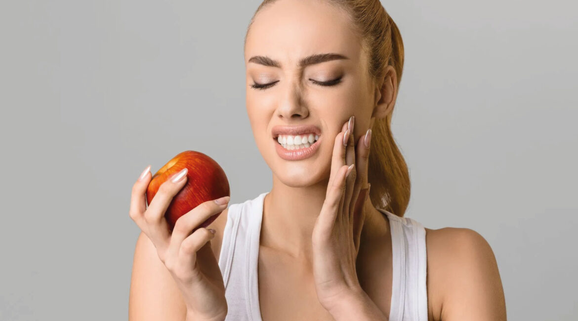 A woman holding an apple and experiencing tooth pain, representing the causes and solutions for sensitive teeth, addressed by Dr. Lenia's Dental Surgery.